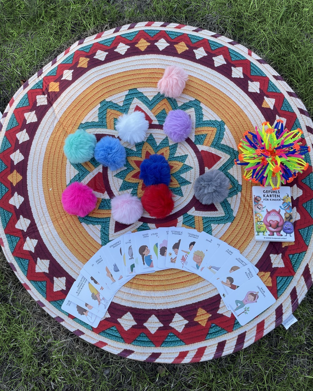 Runder Teppich im Gras mit bunten Pompoms, aufgefächerten Yoga-Karten, Gefühlskarten für Kinder und einem Atemball